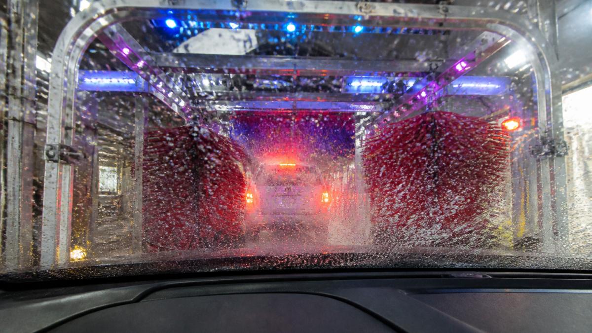 A car moving through a brightly lit express tunnel wash, with colorful soap foam on the windshield and spinning brushes visible ahead, water spray catching the light