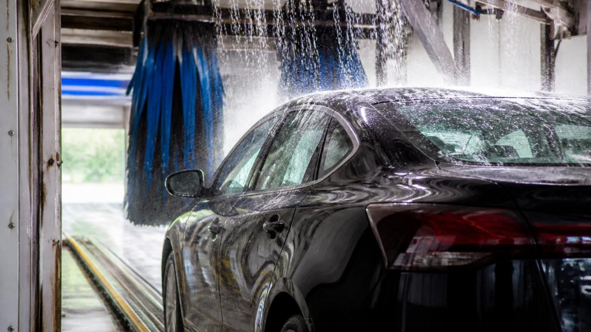 Car entering a brightly lit express car wash tunnel with water spraying and soft brushes cleaning the vehicle.